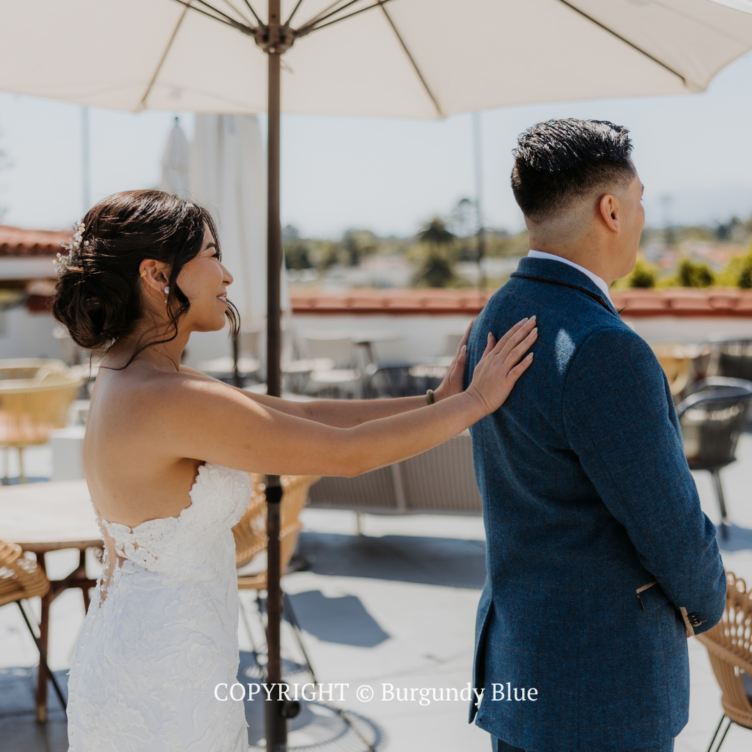Bride in white dress touches groom’s shoulder under an umbrella on a sunny outdoor terrace