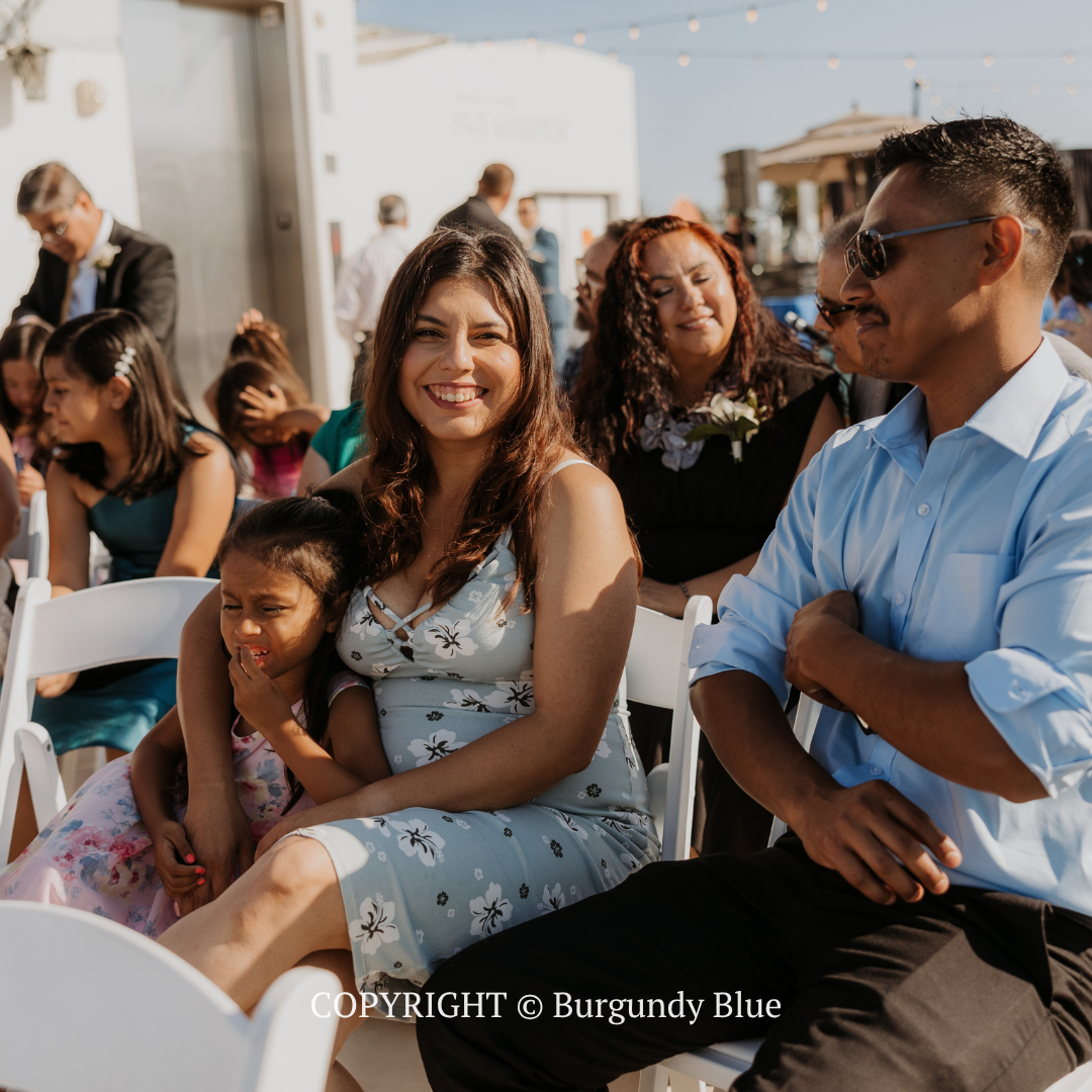 Smiling guests seated outdoors at a sunny event, with a woman holding a child in the front row.