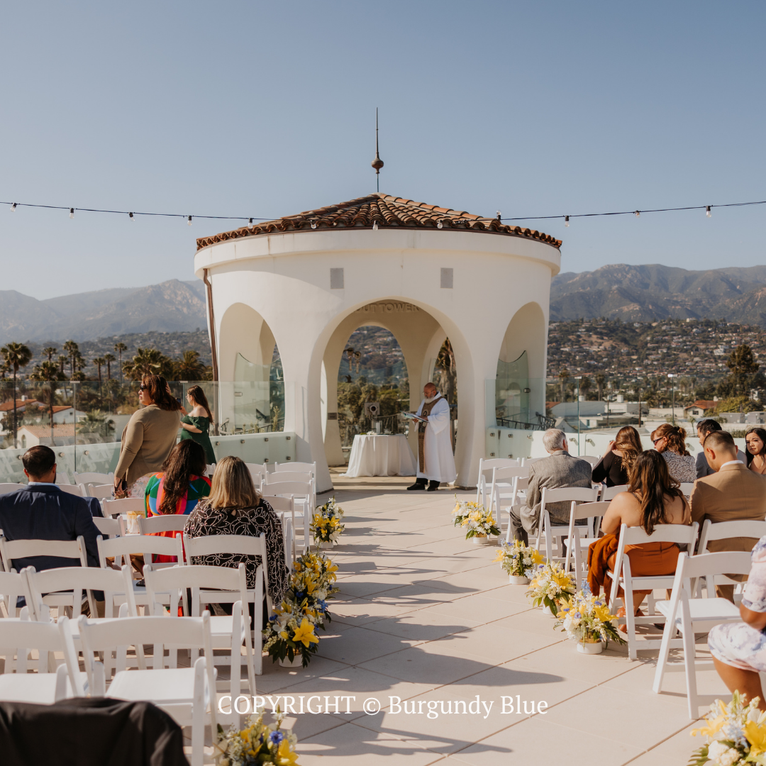 Outdoor wedding ceremony at a white pavilion with guests seated on both sides of a flower-lined aisle.