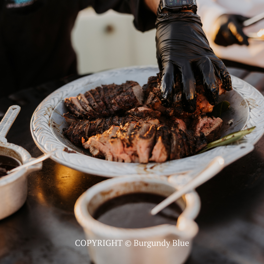 Gloved hand serving sliced grilled steak on a white platter with sauce bowls nearby
