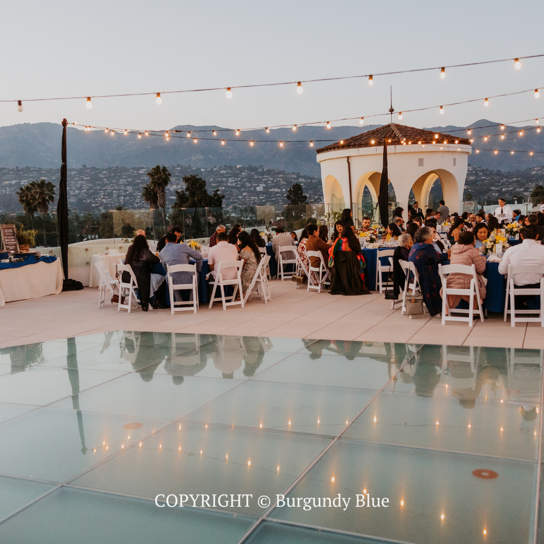 Outdoor evening wedding reception on a terrace with string lights, seated guests, and a city view beyond the pool