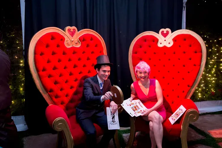 Two people seated in oversized red heart chairs, posing with props in a festive photo booth