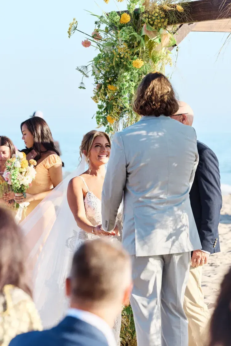 Bride and groom standing under a flower-covered arch at a beach wedding ceremony