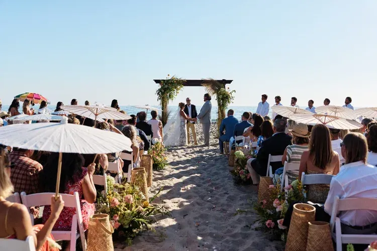 Beach wedding ceremony on a sandy aisle, with guests seated under white umbrellas facing an arch by the sea.