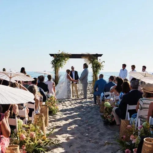 Beach wedding ceremony under a floral arch with guests seated along a sandy aisle