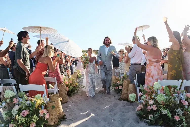 Guests celebrate an outdoor wedding aisle lined with flowers and white umbrellas.