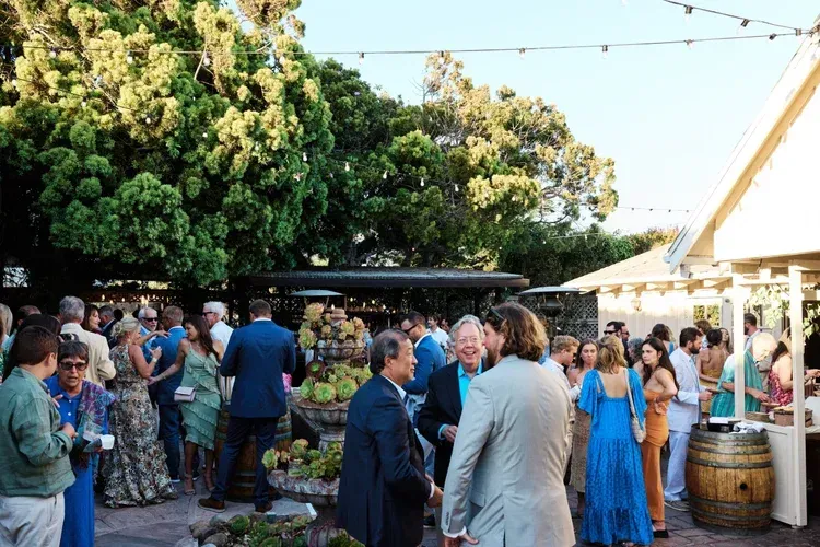 People mingling at an outdoor garden reception with string lights and a white tent beside a large tree.