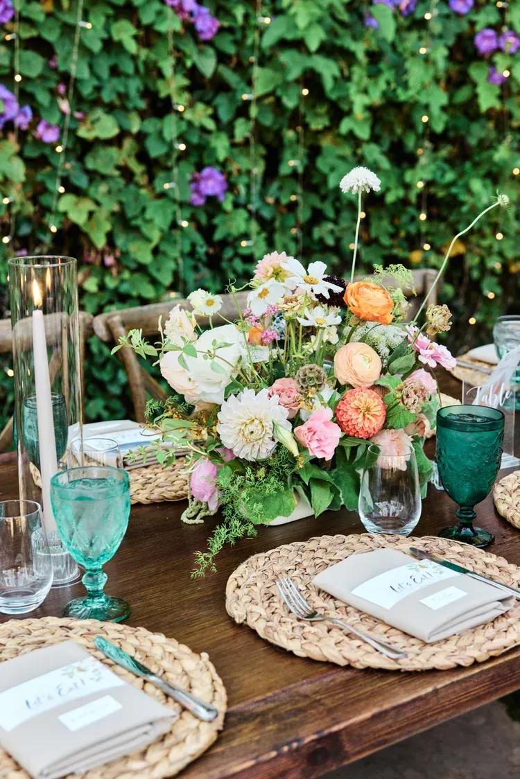 Outdoor table with pastel floral centerpiece, blue glassware, and woven placemats in a garden setting