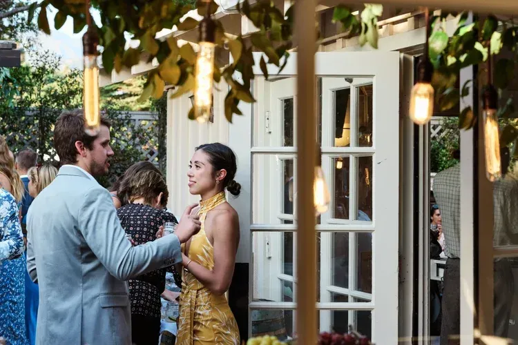 Three people chatting under a leafy canopy outside a doorway, with warm hanging lights and one woman in a gold dress.