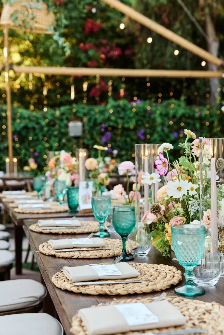 Outdoor banquet table with teal glasses, white napkins, and floral centerpieces under string lights