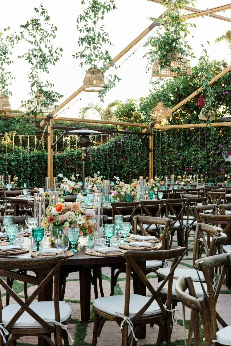 Outdoor dining setup with wooden tables and chairs under a glass-roofed, greenery-filled pavilion