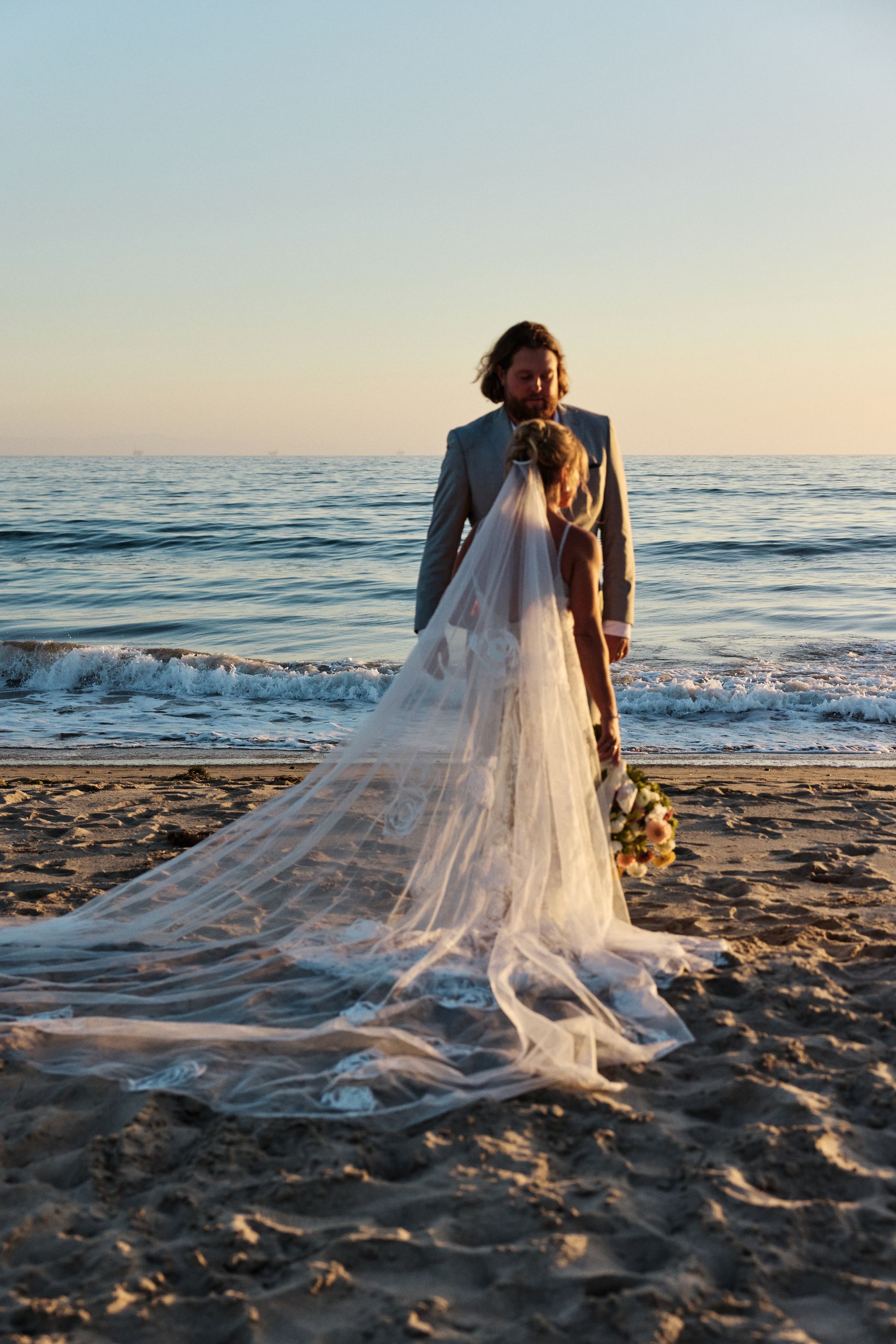 Person in a long flowing veil stands on a beach at sunset, with ocean waves behind.
