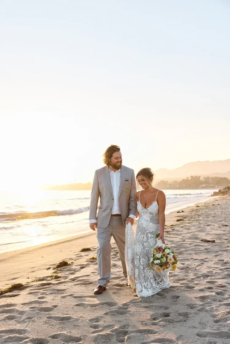 Couple in wedding attire walking barefoot along a beach at sunset, bride holding a bouquet.