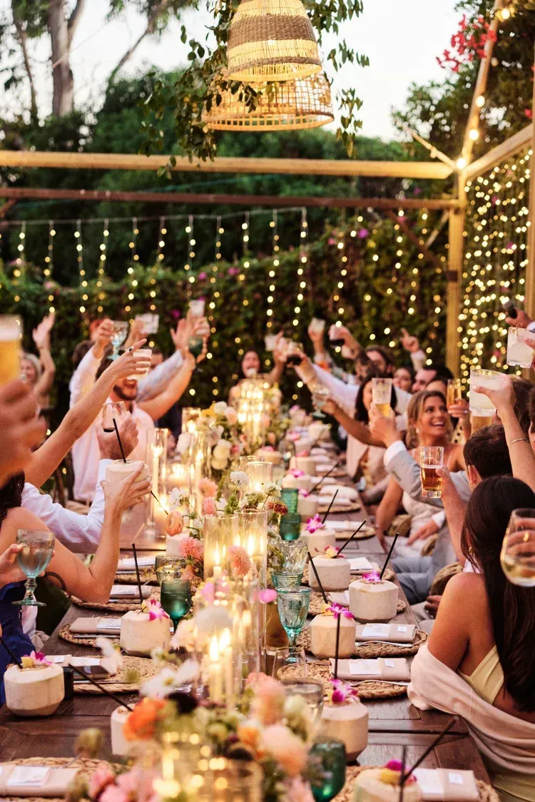 Outdoor dinner party with guests raising glasses at a long candlelit table under string lights