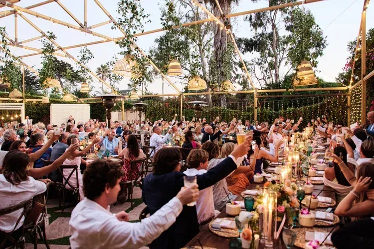 Outdoor banquet with guests raising glasses at long candlelit tables under a floral canopy