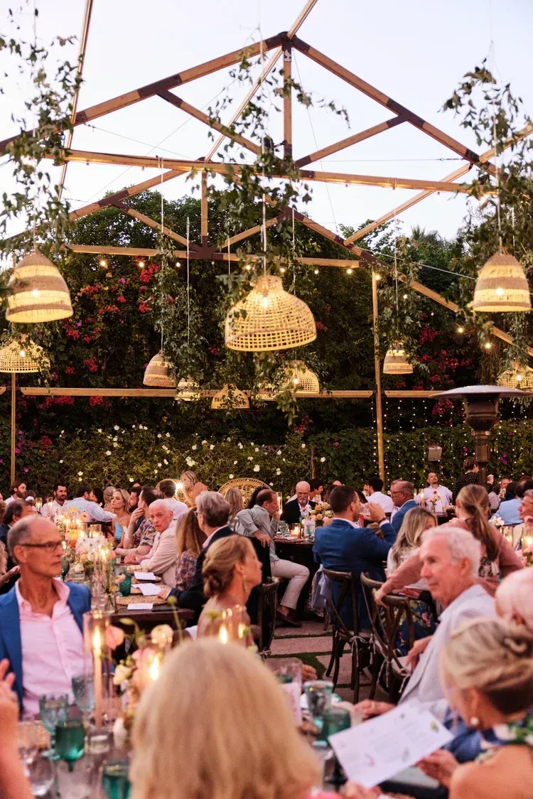 Outdoor banquet with seated guests under hanging lanterns and wooden beams, surrounded by greenery