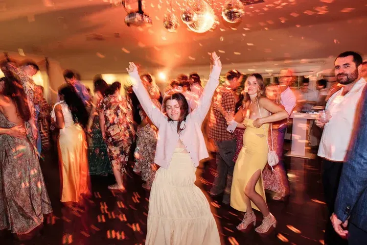 People dancing at a lively indoor celebration, with one person in a cream dress raising both arms.