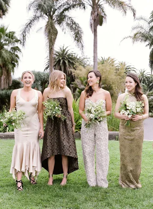 Four women standing outdoors in dresses, holding bouquets, with palm trees in the background.