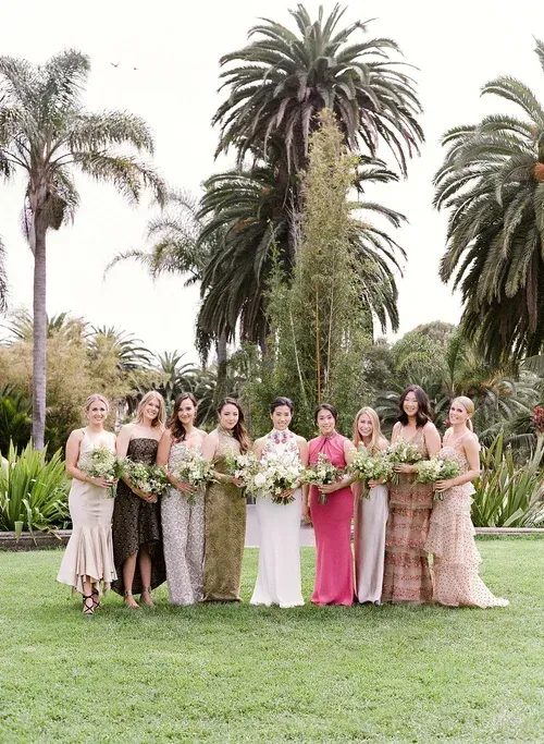 Nine women in pastel dresses holding bouquets pose on a lawn with tall palm trees behind them.