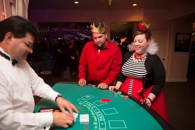 People playing casino roulette at a green table, with a dealer on the left and two players wearing festive crowns.