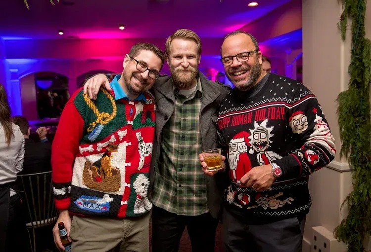 Three smiling friends in festive sweaters posing at a colorful party, one holding a drink