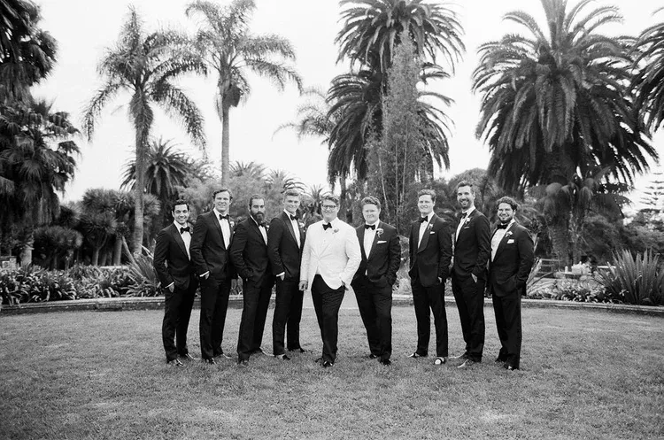 Group of eight people in formal suits standing outdoors on a lawn with palm trees behind them