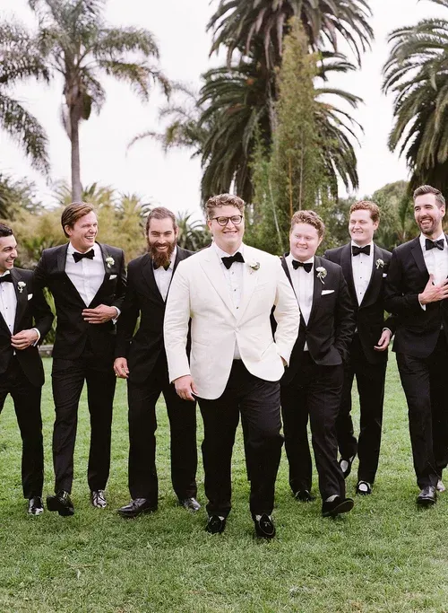 Group of men in tuxedos and a white suit posing on a lawn with palm trees behind them