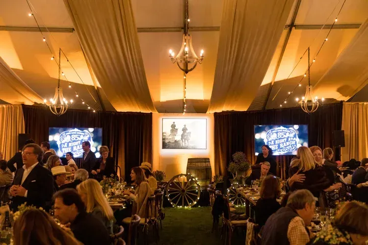 Elegant indoor banquet with seated guests, gold drapes, chandeliers, and large projection screens at the front