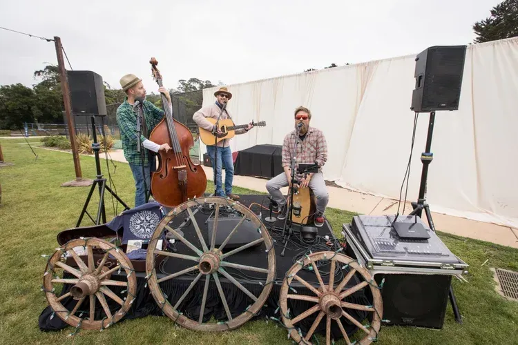 Three musicians performing on a small outdoor stage with instruments and wagon wheels in front
