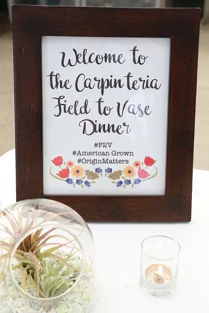 Framed sign reading “Welcome to the Carpinteria Field to Table Dinner” with flowers and a small plant beside it