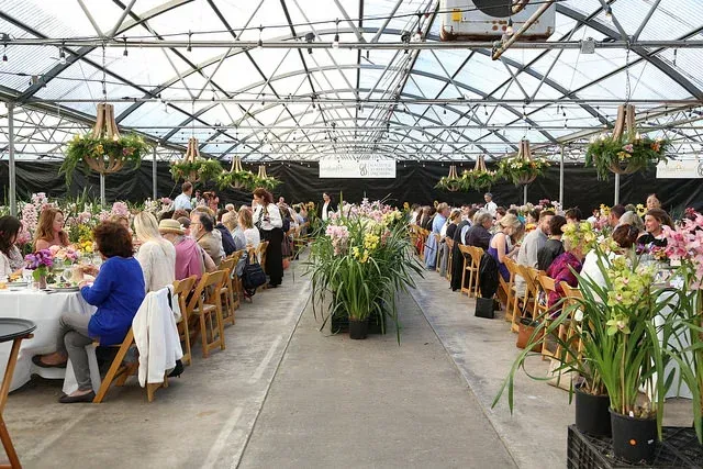 Crowded indoor banquet in a greenhouse, with guests seated at long tables under a glass roof surrounded by plants