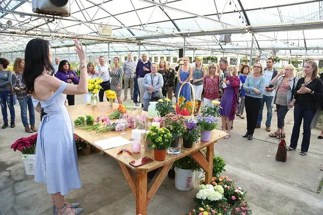 Flowers displayed on a table as a woman speaks to a gathered crowd in a greenhouse