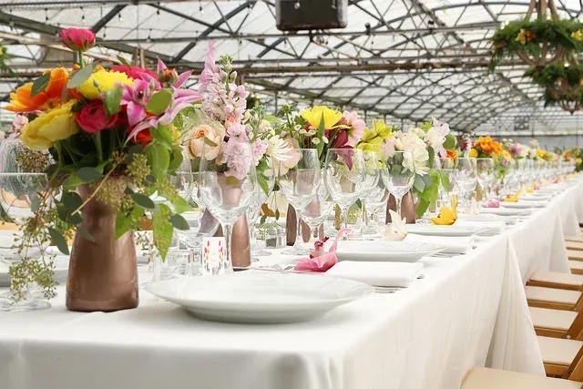 Long banquet table in a greenhouse, set with white plates and colorful floral centerpieces.