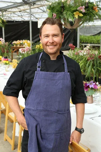 Smiling man in a blue apron standing in a flower shop greenhouse with hanging and potted flowers