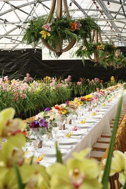 Long banquet table in a flower-filled greenhouse, decorated with colorful blooms and hanging baskets