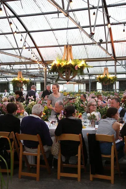 Guests seated at a banquet table in a glass greenhouse, with hanging flower arrangements overhead