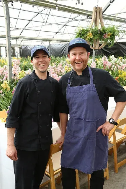 Two smiling greenhouse workers in black and blue aprons standing among hanging plants