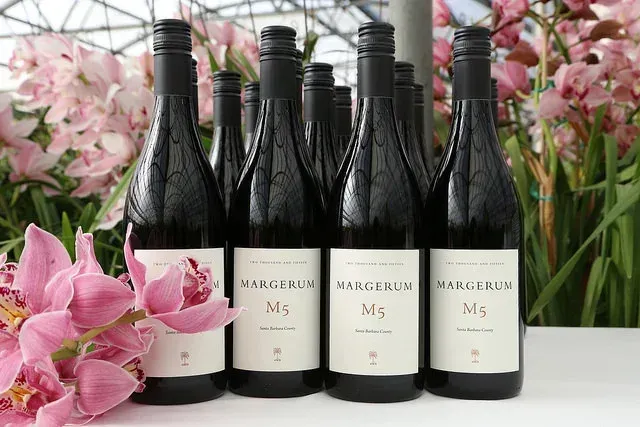 Five black wine bottles with white labels arranged among pink flowers in a greenhouse