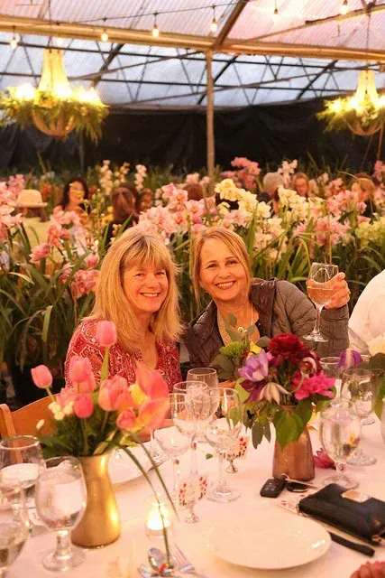 Two women smiling at a candlelit flower-filled dinner table under a glass-roofed tent