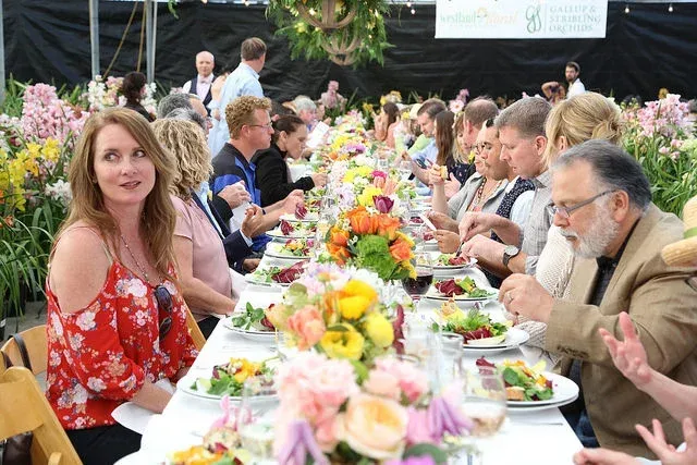 Outdoor banquet with guests seated at a long flower-decorated table, eating and talking