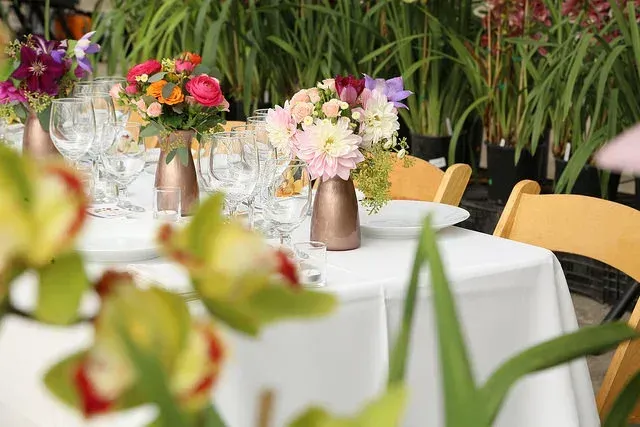 Outdoor banquet table with white tablecloth, floral centerpieces, glassware, and green plants in the background