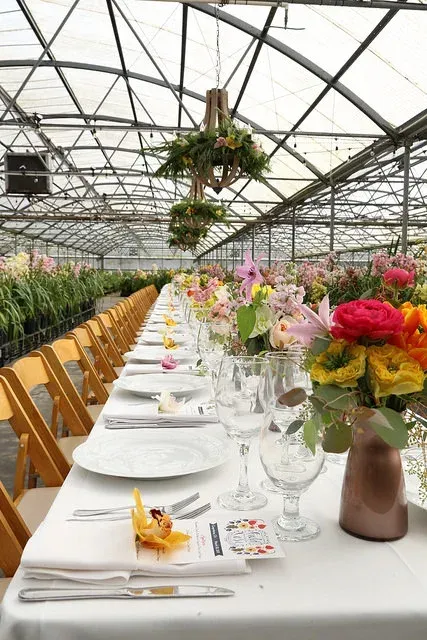 Long banquet table in a greenhouse, set with white linens and colorful flowers under a glass roof
