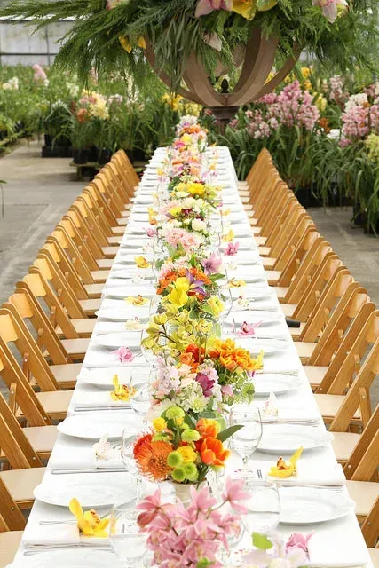 Long banquet table with white tablecloth, floral centerpieces, and wooden chairs in a lush garden setting