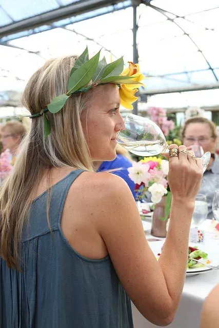 Woman in a blue dress and flower crown sips wine at an outdoor table with floral centerpieces.