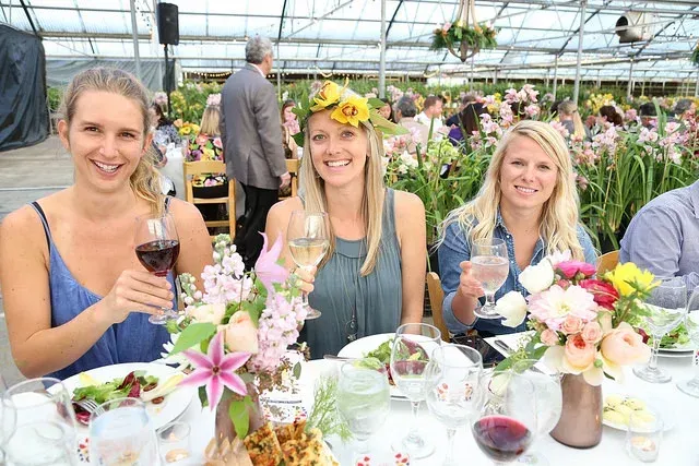 Three women smiling at a flower-filled banquet table in a greenhouse, one holding a wine glass