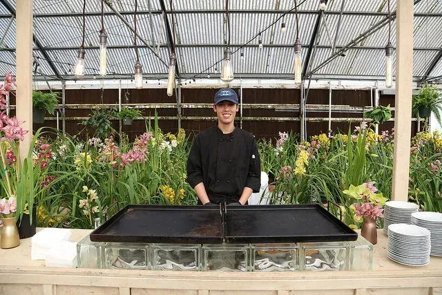 Person standing behind black trays in a greenhouse with flowers and plants in the background
