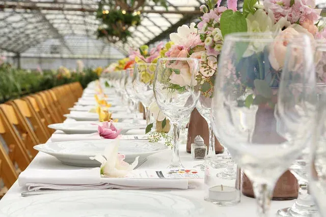 Elegant long banquet table set with white tableware and floral centerpieces in a bright greenhouse