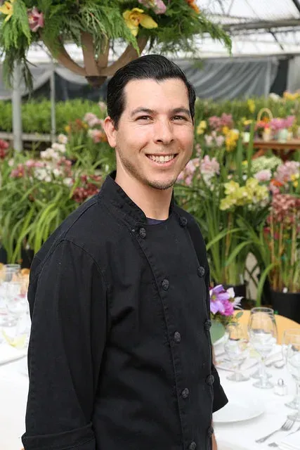 Smiling man in a black shirt standing at a table in a flower-filled event space