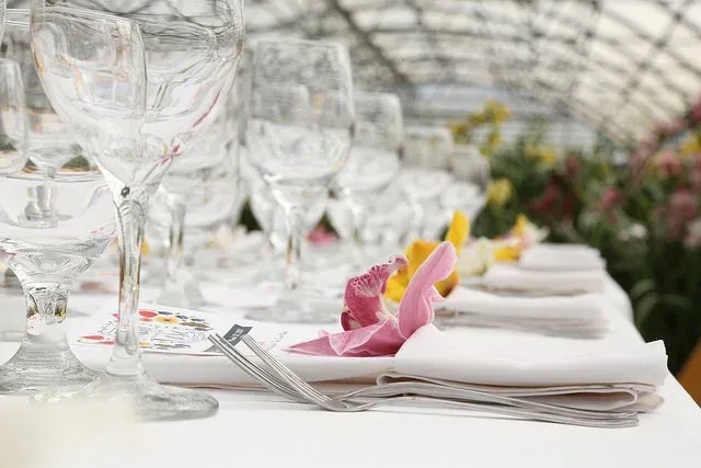Elegant banquet table with white linens, glassware, folded napkins, and pink flower petals in a bright event hall
