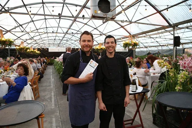 Two chefs posing in a greenhouse dining hall with seated guests and tables in the background